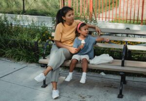 Woman with Brown skin and dark hair sitting on a bench beside a child with Brown skin and dark hair holding an ice cream cone